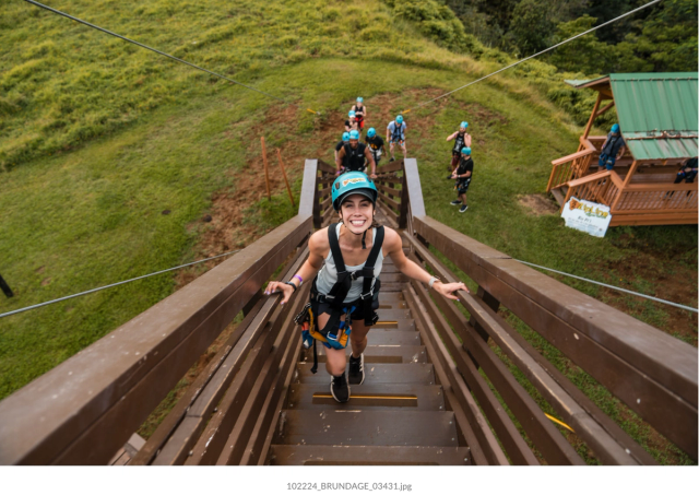 A happy woman in zip-lining gear climbs wooden stairs, with other people and a green landscape in the background.