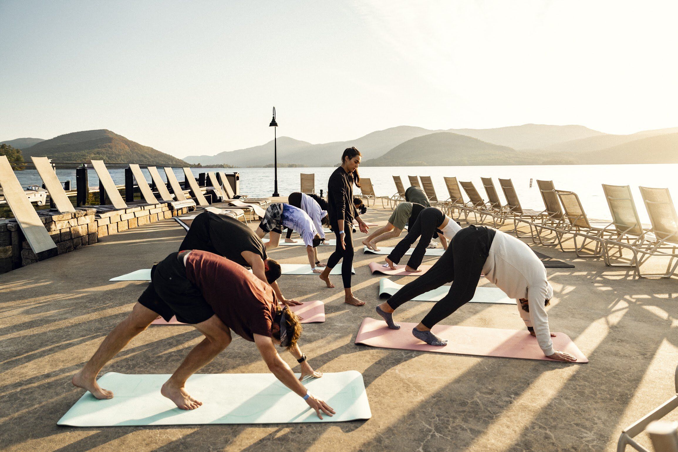 A group practices yoga in downward dog pose outdoors by a lake with mountains in the distance.