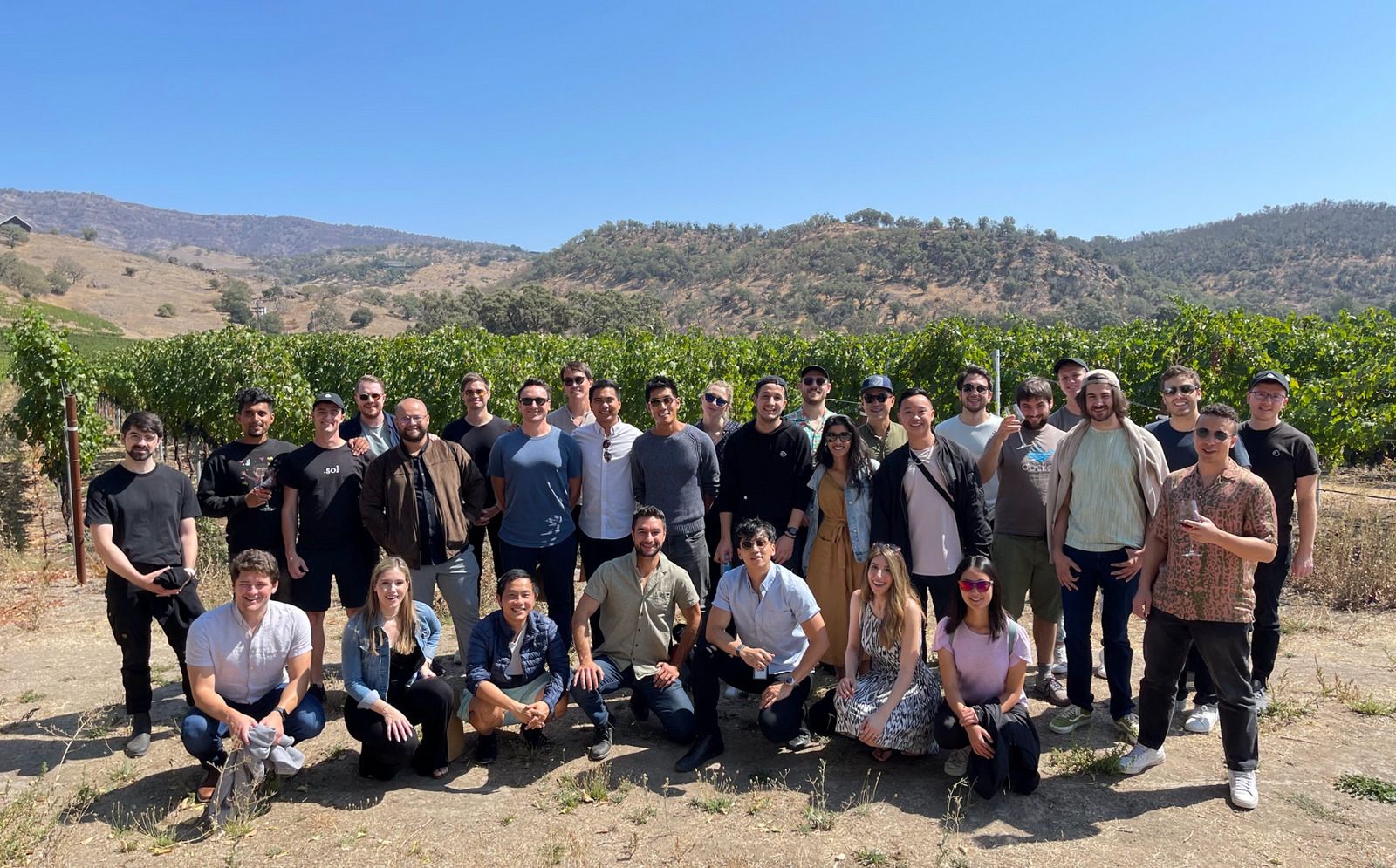 A large group of people posing in a vineyard with hills in the background.