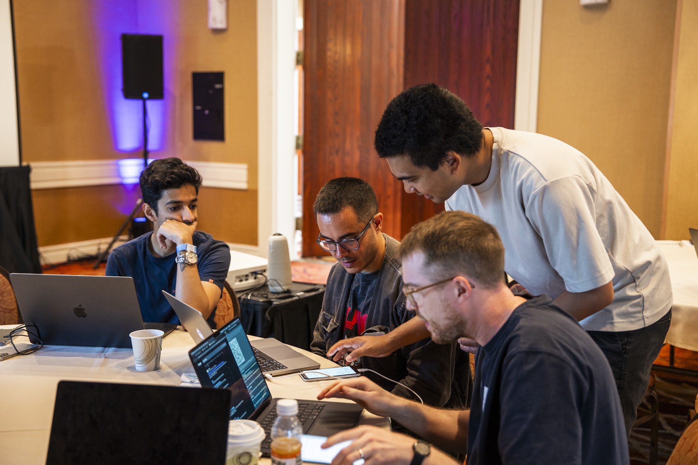 Four men collaborarate at a table with laptops, with one standing and leaning over to assist another.