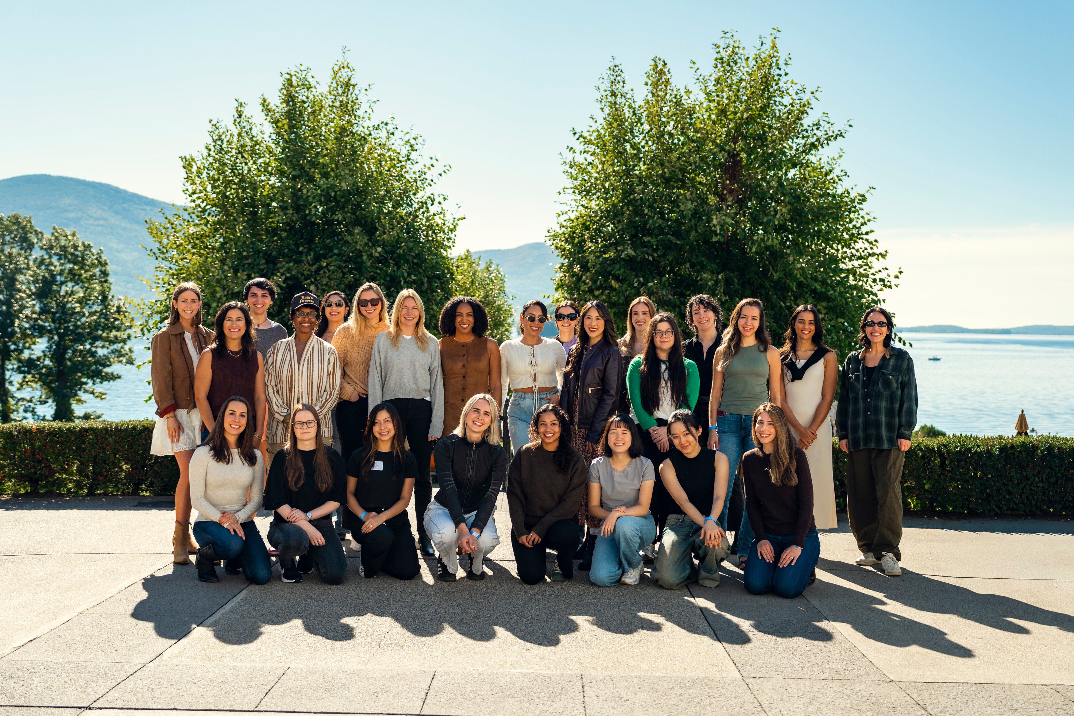 A group of diverse young adults, predominantly women, smiles while posing outdoors against a scenic backdrop of water, mountains, and trees.